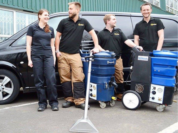 This is a photo of Haggerston Carpet Cleaning carpet cleaners (three men and one woman) standing in fromt of their black van, with two steam cleaning carpet machines next to them